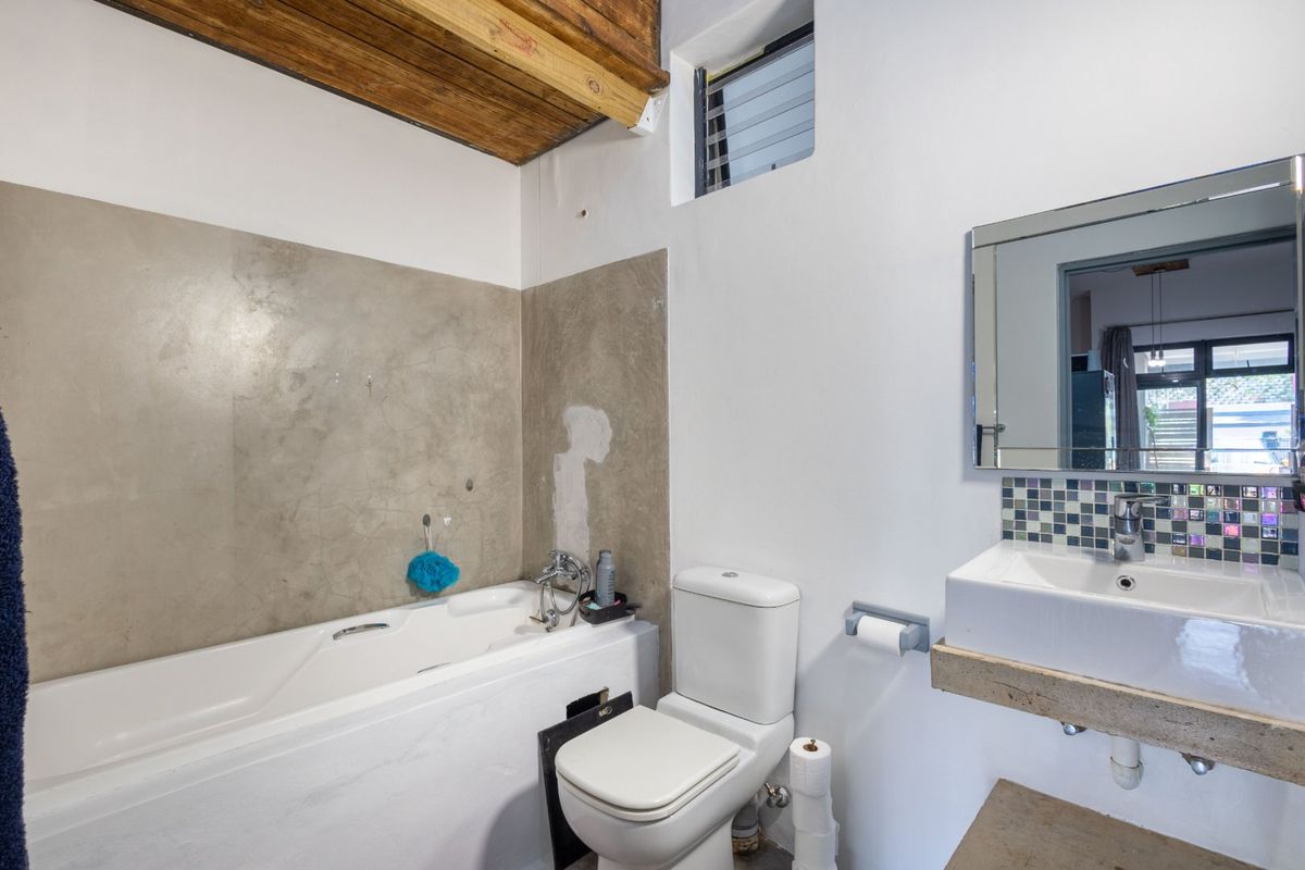 A compact bathroom featuring a bathtub, wall-mounted basin, and toilet, with exposed timber ceiling beams and unfinished wall plastering requiring cosmetic attention.