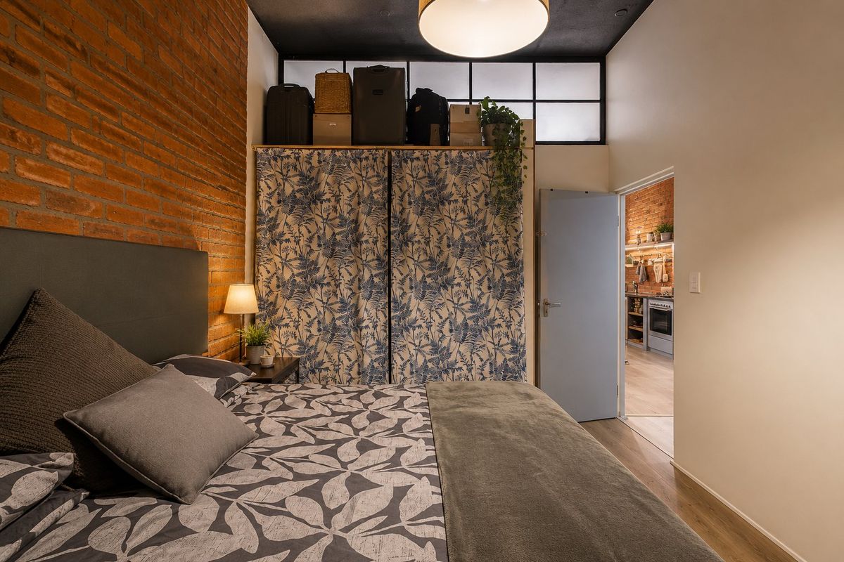 A cozy bedroom featuring an exposed brick wall, high ceilings with a transom window, and built-in storage, with a view into the adjacent kitchen.