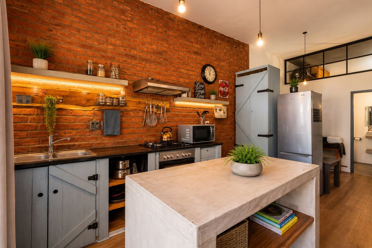Modern industrial-style kitchen featuring an exposed brick wall, concrete island, timber shelving, and high-quality built-in appliances with warm ambient lighting.