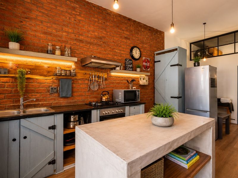 Modern industrial-style kitchen featuring an exposed brick wall, concrete island, timber shelving, and high-quality built-in appliances with warm ambient lighting.