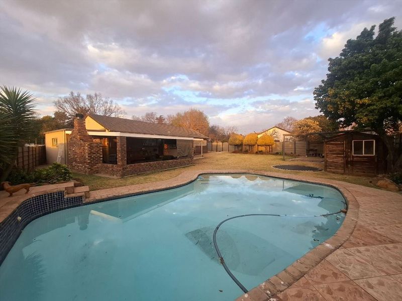 Spacious backyard featuring a swimming pool, covered patio area with brick chimney, and a timber storage shed set within a landscaped garden.