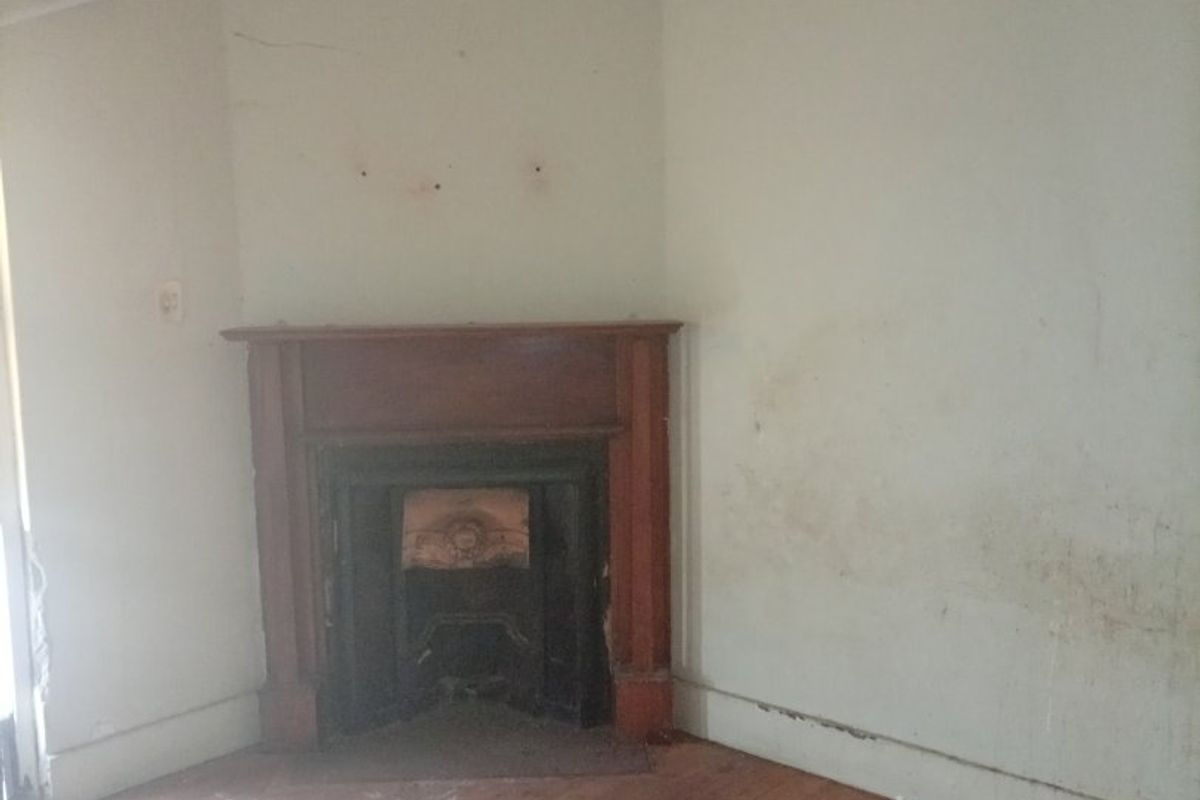 An empty living room featuring original timber floorboards and a classic cast iron fireplace with a timber mantelpiece, requiring cosmetic refurbishment.