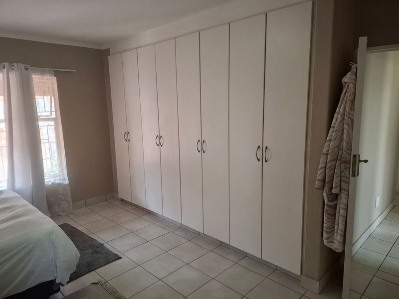 A bedroom featuring extensive floor-to-ceiling built-in cupboards, neutral tiled flooring, and a window with security bars.
