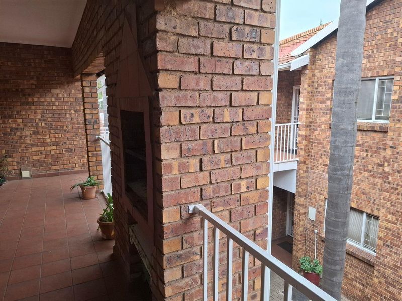 A covered balcony featuring a built-in brick braai, tiled flooring, and metal railings, overlooking the townhouse complex courtyard.