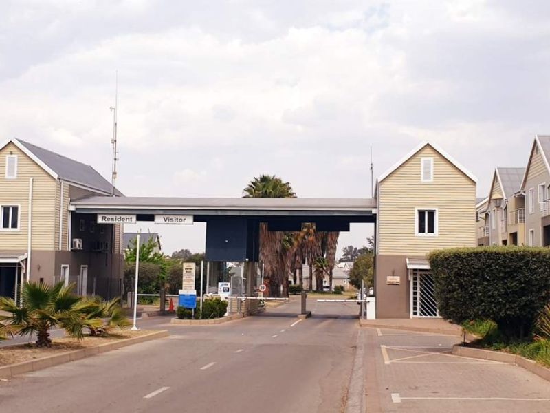 The main entrance to the apartment complex featuring a secure gatehouse, boom gates for residents and visitors, and well-maintained paved access roads.