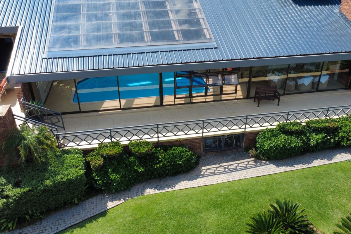 Aerial view of a property featuring a swimming pool, a large skylight on a metal roof, a tiled patio, and a well-maintained garden.