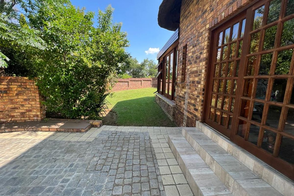 Rear exterior view featuring a paved patio, face brick facade, thatched roof detail, and timber-framed glass doors leading to a well-maintained garden.