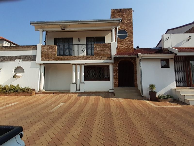 Front elevation of a multi-level house featuring a paved driveway, brick facade, balcony, and secure entrance with decorative columns.