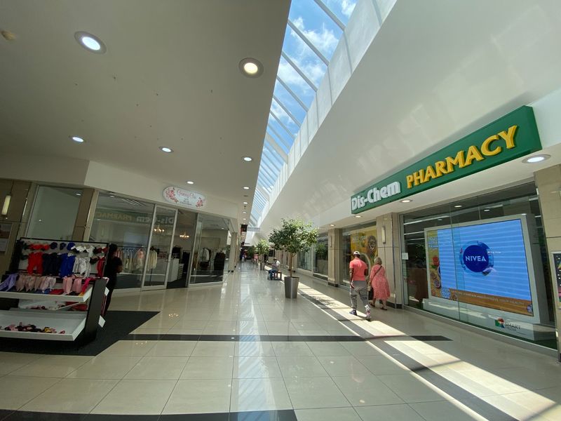 A bright, modern commercial shopping centre corridor featuring a large skylight, polished tiled floors, and glass-fronted retail units.