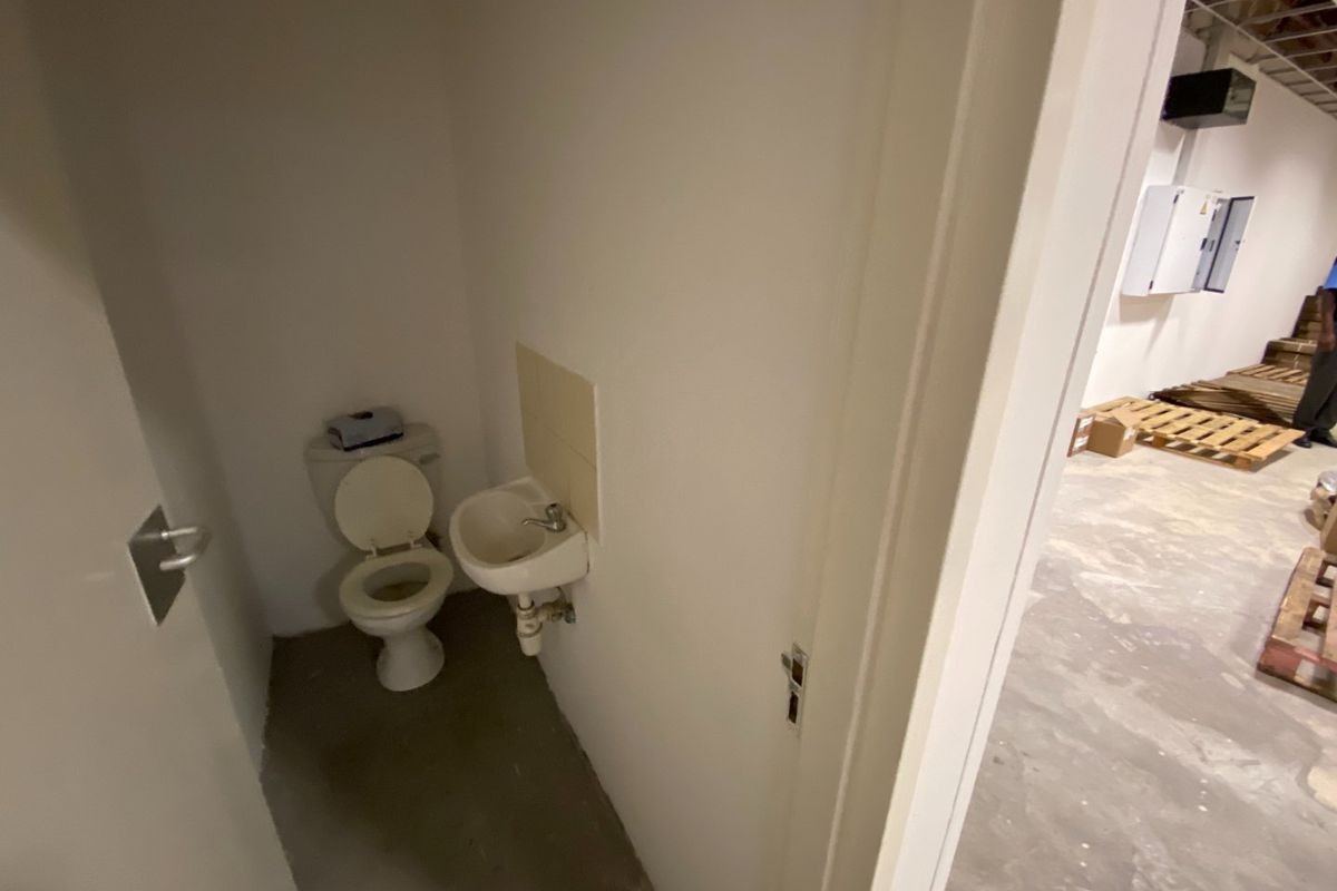 A small, functional commercial restroom featuring a toilet and wall-mounted basin, located adjacent to an unfinished utility or storage area.