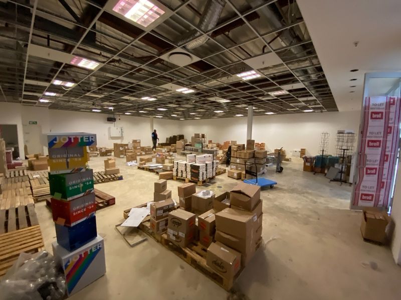Large open-plan commercial space featuring a suspended ceiling grid with missing tiles, concrete flooring, and support columns, currently utilized for storage.