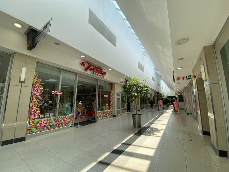 A bright, modern commercial shopping centre corridor featuring tiled floors, a high skylight ceiling, and glass-fronted retail units.