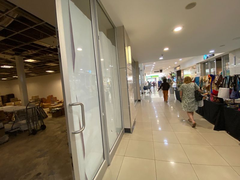 A commercial hallway view showing a vacant retail unit with exposed ceiling grid alongside an active shopping corridor with tiled flooring and recessed lighting.