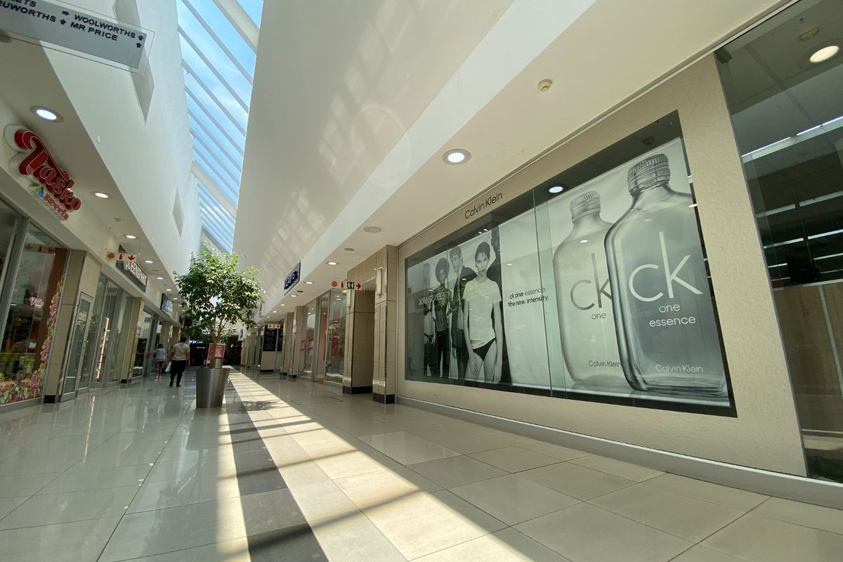 A bright, modern commercial shopping mall corridor featuring high ceilings with skylights, polished tiled floors, and glass-fronted retail storefronts.