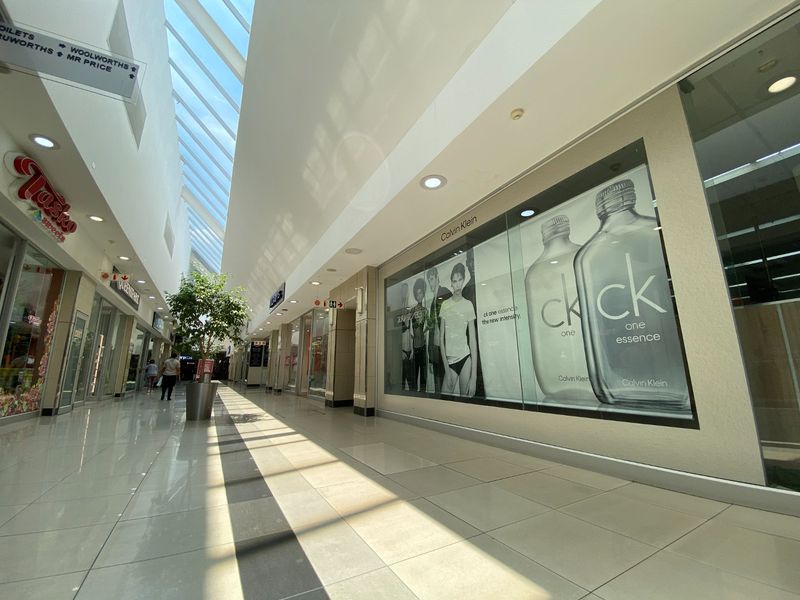 A bright, modern commercial shopping mall corridor featuring high ceilings with skylights, polished tiled floors, and glass-fronted retail storefronts.