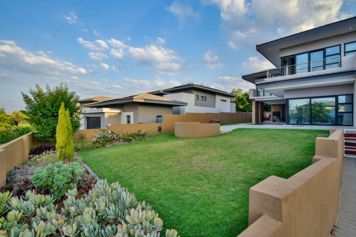 A well-maintained rear garden featuring a manicured lawn, landscaped flower beds, boundary walls, and a modern multi-story home with a balcony and patio area.