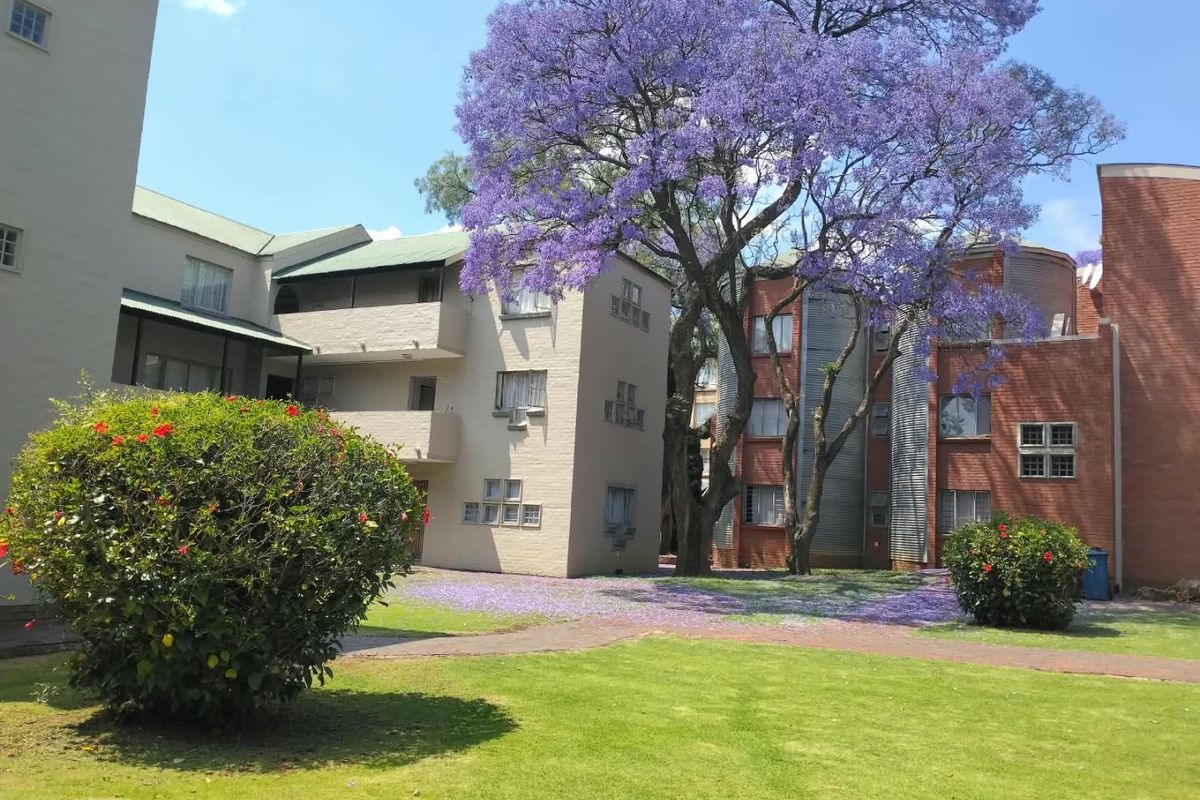 Exterior view of the apartment complex featuring brick architecture, balconies, and a well-maintained garden with a large flowering jacaranda tree.