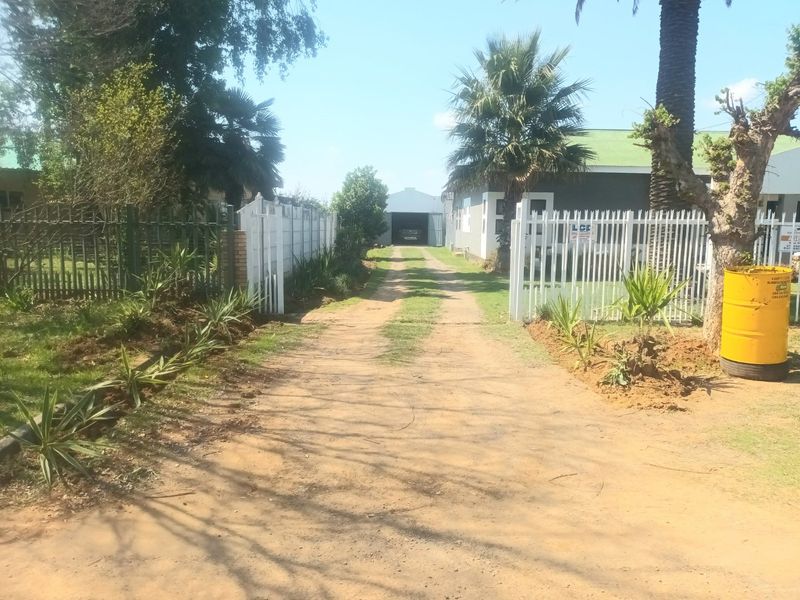 A view of the property entrance featuring a long unpaved driveway, metal security fencing, and a detached garage structure at the rear.