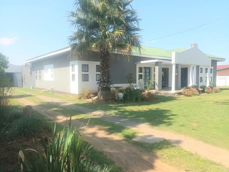 A single-story residential house featuring a covered front porch with decorative columns, a metal roof, and a large front garden with a driveway.