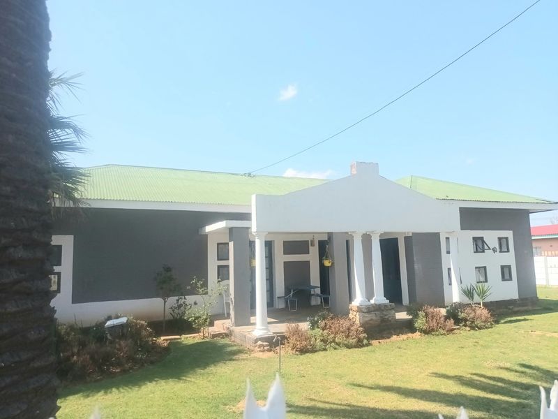 Front elevation of a single-story house featuring a covered porch with classical columns, a green corrugated metal roof, and a well-maintained front lawn.