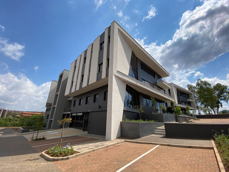 Modern commercial building exterior featuring contemporary architectural design, large glass windows, paved parking area, and landscaped garden beds.