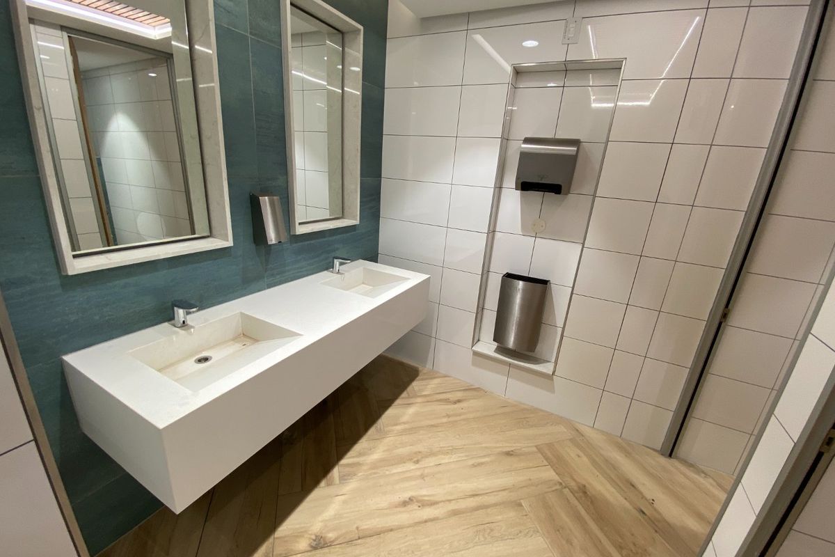 Modern commercial bathroom featuring a sleek double vanity, recessed wall mirrors, integrated hand dryer, and stylish herringbone wood-look tile flooring.