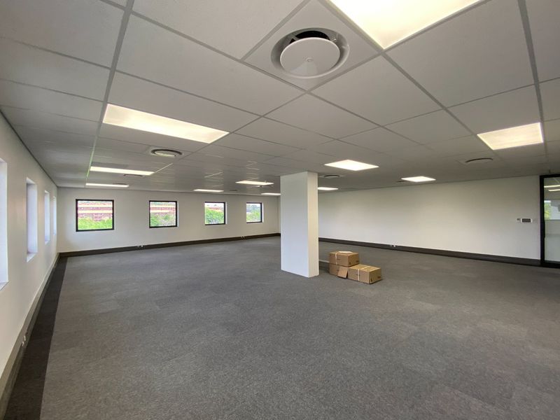 A spacious, open-plan commercial office area featuring neutral carpet tiles, a suspended ceiling with recessed lighting, and central air conditioning.