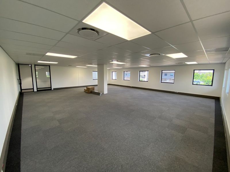 A spacious, open-plan commercial office area featuring neutral carpet tiles, a suspended ceiling with recessed lighting, and large windows providing natural light.
