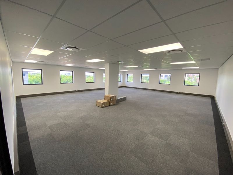 A spacious, open-plan commercial office area featuring neutral carpet tiles, a suspended ceiling with recessed lighting, and large windows providing natural light.