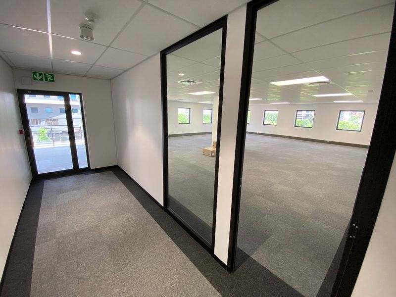 A bright, open-plan commercial office space featuring modern glass partitions, carpeted flooring, and a suspended ceiling with recessed lighting.