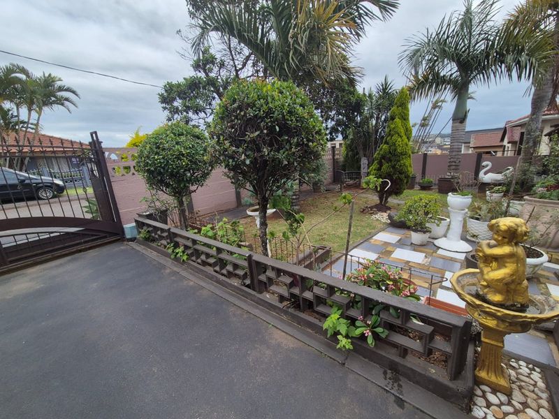 A landscaped garden area featuring a paved driveway, metal gate, boundary wall, and a tiled patio section with established trees and shrubs.