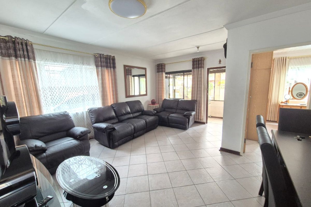 A spacious living room featuring neutral tiled flooring, large windows with curtains, and direct access to an adjacent room through a doorway.