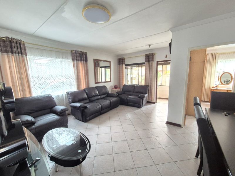 A spacious living room featuring neutral tiled flooring, large windows with curtains, and direct access to an adjacent room through a doorway.