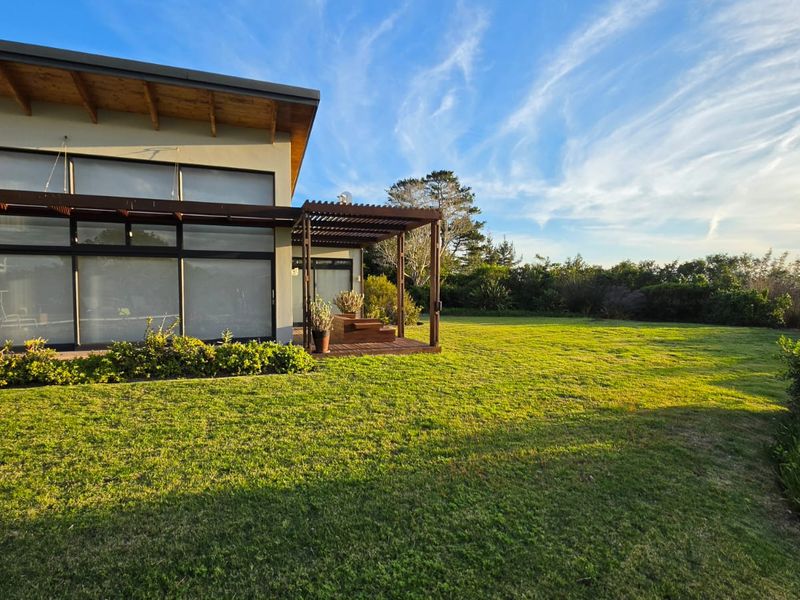 Rear view of a modern farm residence featuring a timber pergola, expansive decking, and large glass sliding doors overlooking a well-maintained lawn.