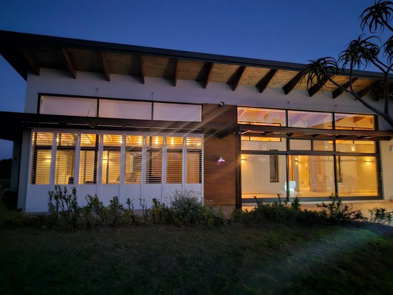 A modern rear facade featuring extensive glass sliding doors, timber ceiling rafters, and integrated timber cladding, opening onto a paved patio area at dusk.
