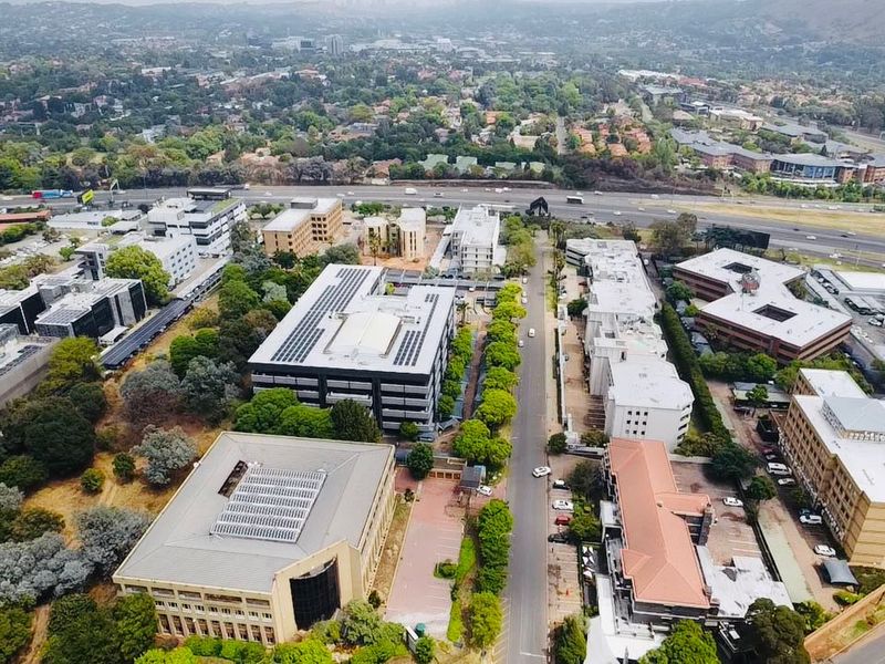 An aerial view of a commercial office park featuring multiple buildings, solar panel installations, paved parking, and surrounding landscaped greenery.