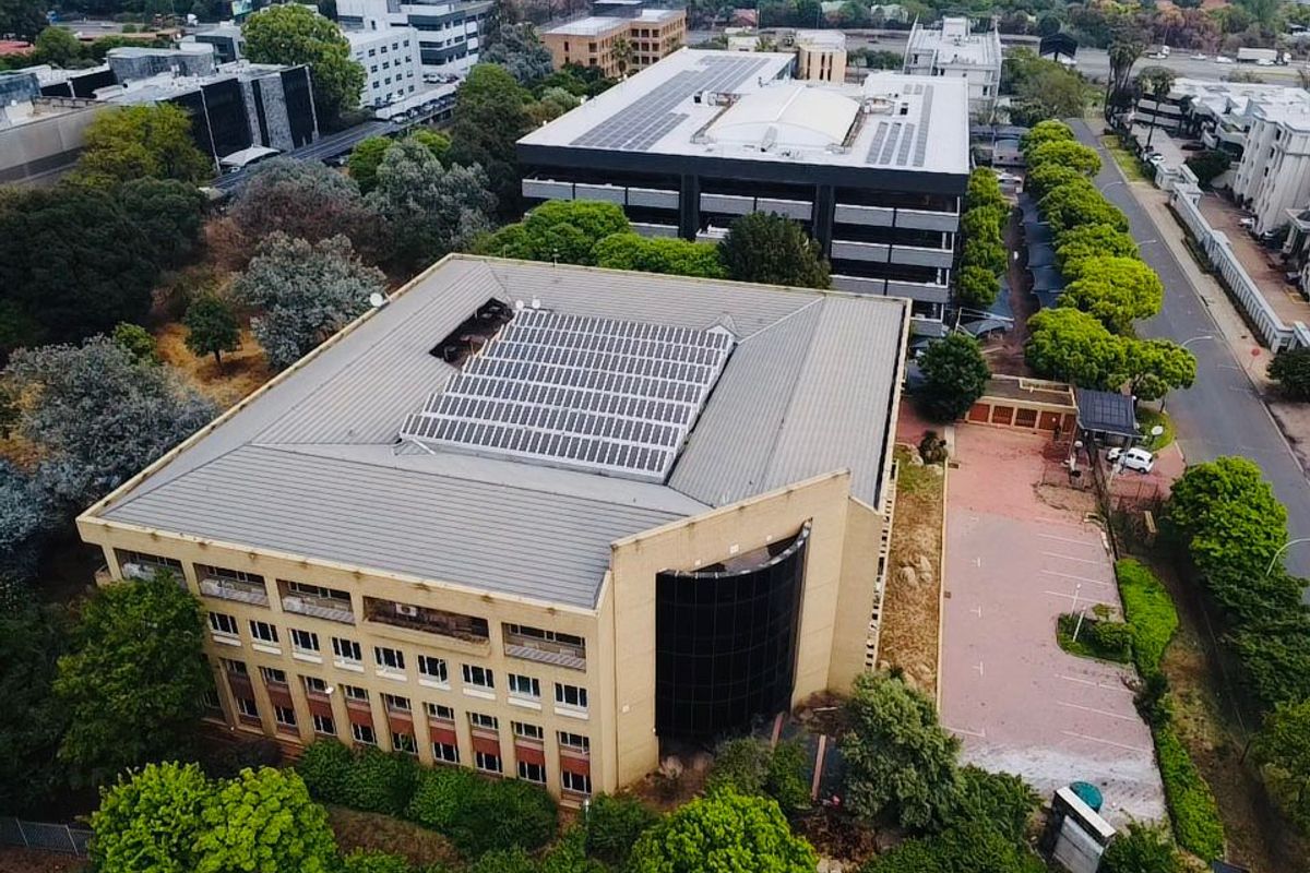Aerial view of a commercial office building featuring a large solar panel array on the roof, paved parking, and surrounding landscaped grounds.