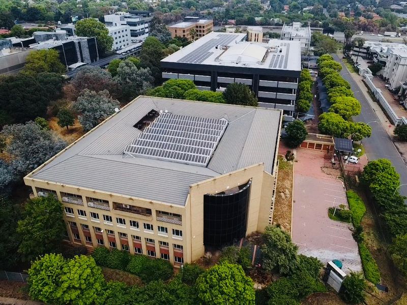 Aerial view of a commercial office building featuring a large solar panel array on the roof, paved parking, and surrounding landscaped grounds.