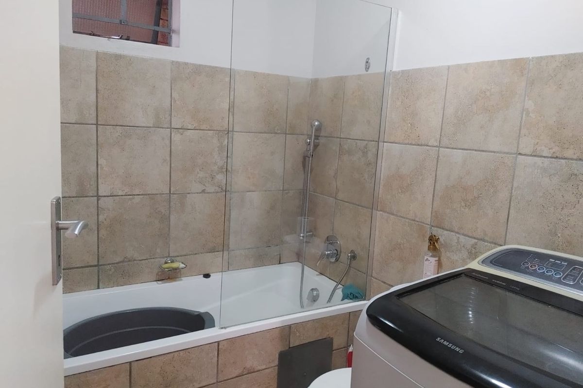 A functional bathroom featuring a bathtub with a glass shower screen, tiled walls, and a window with security bars.