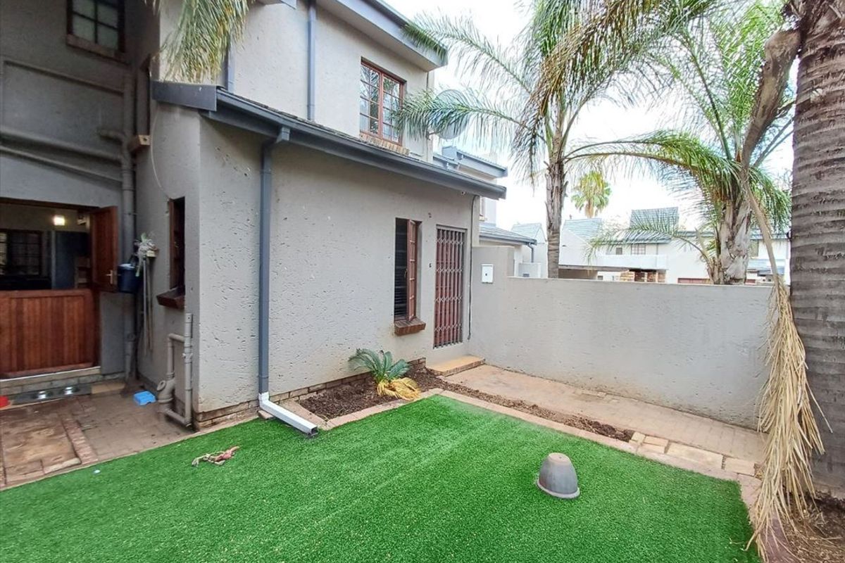 Rear exterior view of the townhouse featuring a small artificial lawn, paved walkway, boundary wall, and direct access to the home via a security gate.