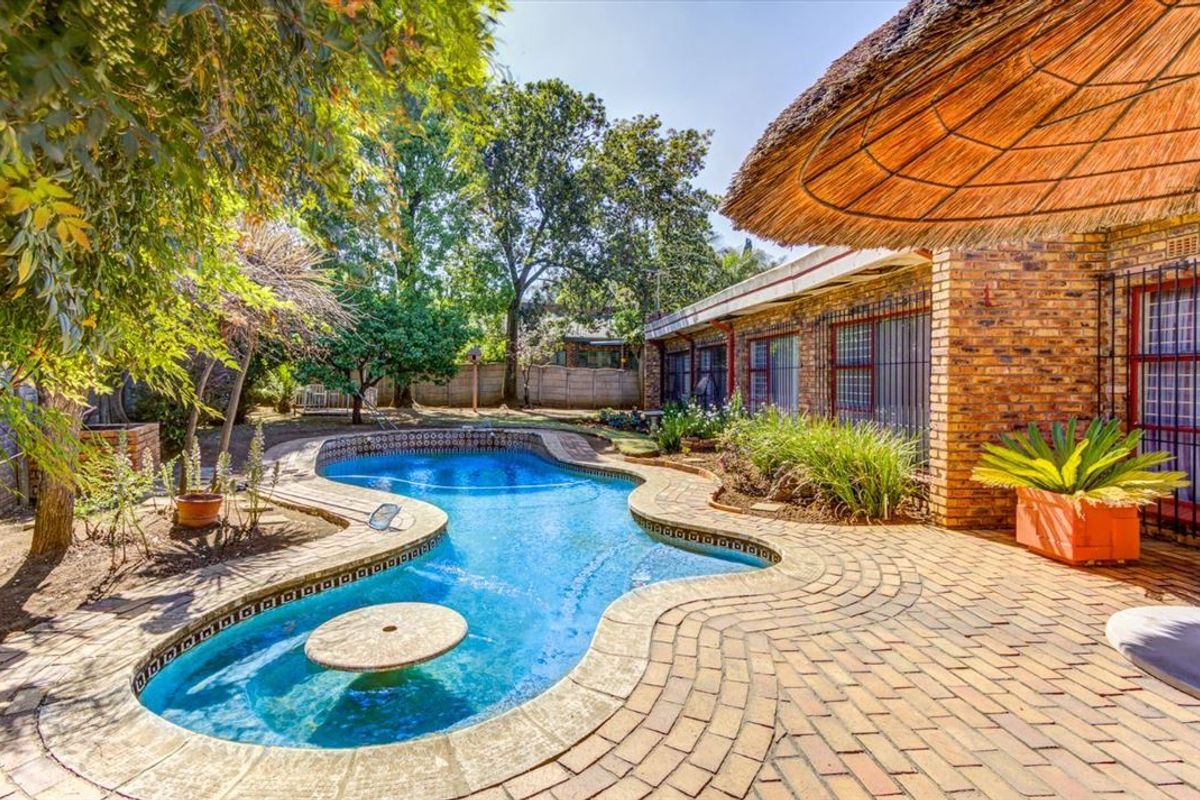 Outdoor pool area featuring a curved swimming pool, brick paved patio, and a thatch umbrella, set within a landscaped garden.