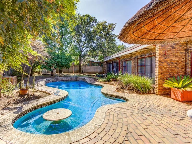 Outdoor pool area featuring a curved swimming pool, brick paved patio, and a thatch umbrella, set within a landscaped garden.