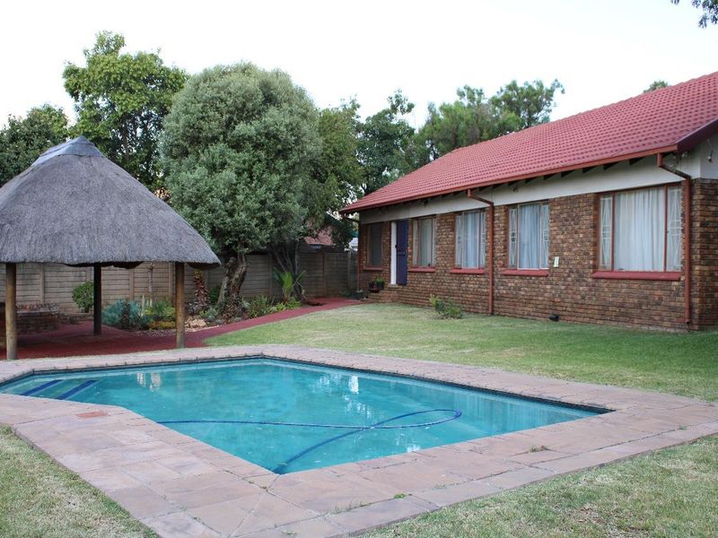 A spacious backyard featuring a swimming pool with paved surround and a traditional thatched lapa, set against a brick-built house with a tiled roof.