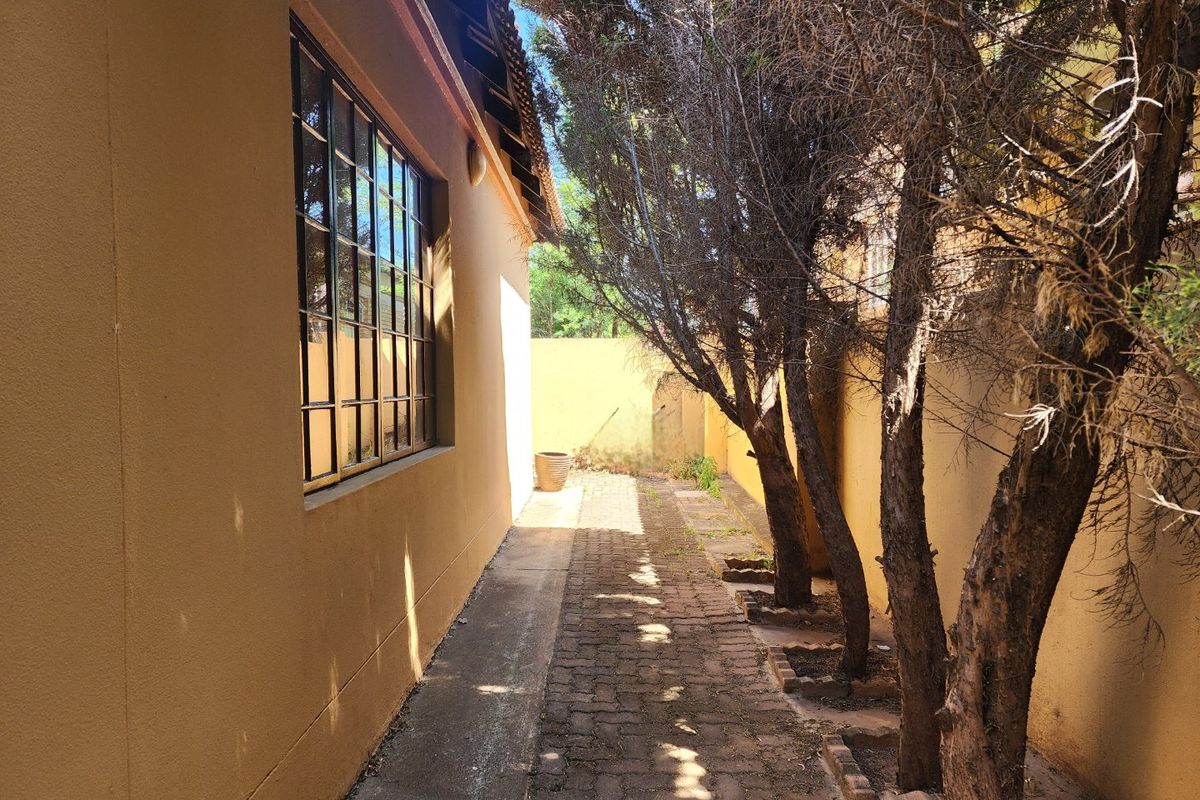 A narrow side passage of the townhouse featuring a brick paved walkway, boundary wall, and mature trees alongside the building exterior.