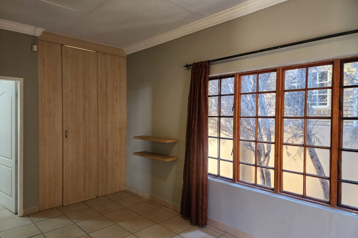 A bright, empty bedroom featuring tiled flooring, built-in wooden cupboards, floating shelves, and large wooden-framed windows with natural light.