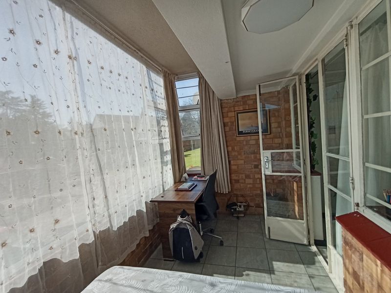 A sun-filled enclosed balcony or patio area featuring exposed brick walls, tiled flooring, and large windows, currently utilized as a home office space.