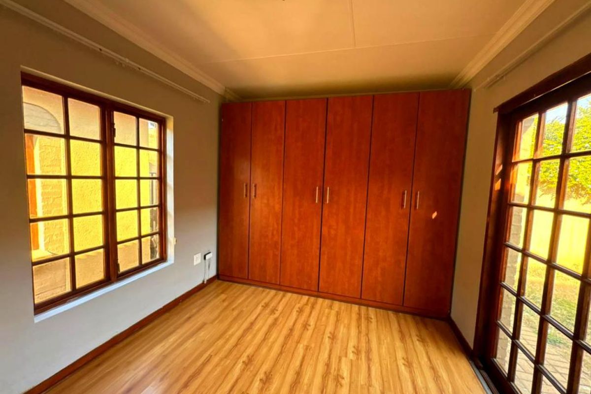 A bright, empty bedroom featuring laminate flooring, built-in wooden cupboards, and large windows that provide ample natural light.