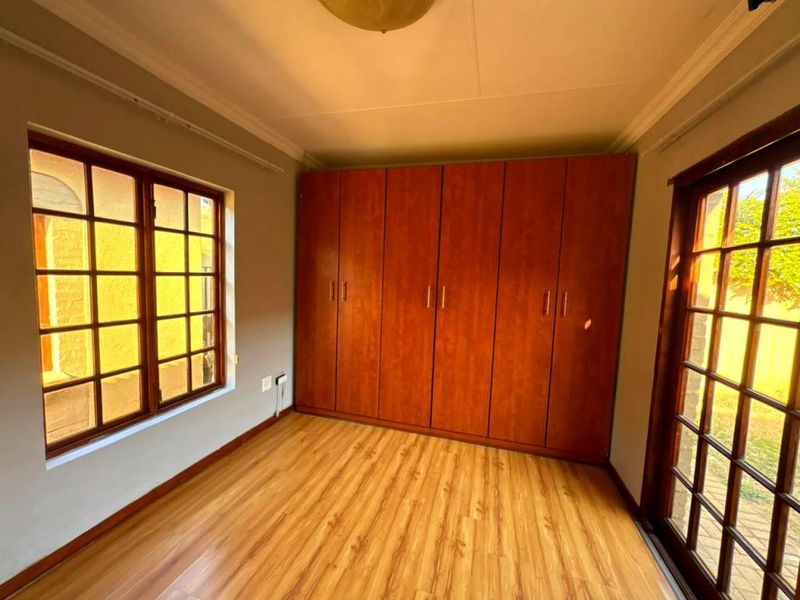 A bright, empty bedroom featuring laminate flooring, built-in wooden cupboards, and large windows that provide ample natural light.