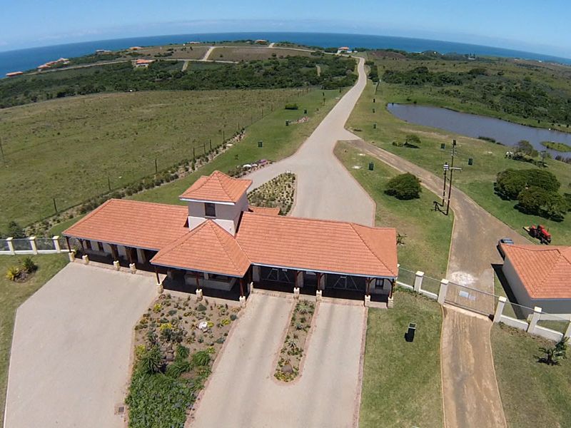Aerial view of a residential estate entrance featuring a gated security house, paved driveway, and landscaped grounds with coastal surroundings.