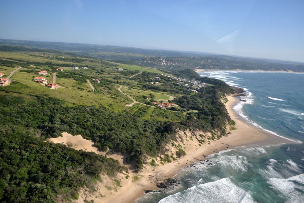An aerial view of a coastal residential development featuring paved roads, natural vegetation, and direct proximity to the ocean and sandy beach.